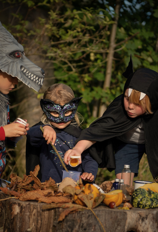 Three children in costumes standing around a potion making kit in a forest setting.