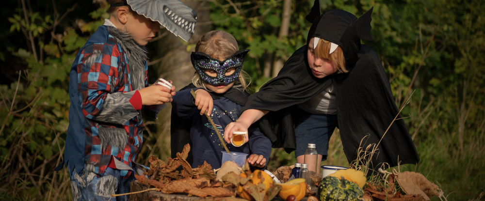 Three children in costumes standing around a potion making kit in a forest setting.