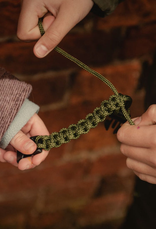 Two hands holding a green knotted cord bracelet against a blurred natural background
